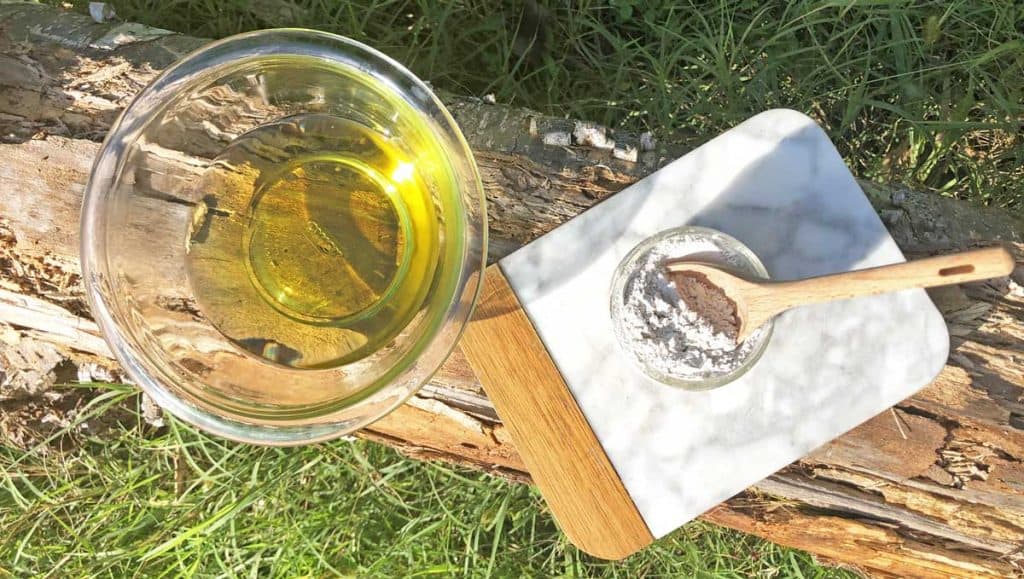 A bowl of melted shea butter, cacao butter, and coconut oil is beside a marble platter with a small bowl of bentonite clay and a wooden tablespoon. These items are outdoors, on a log.