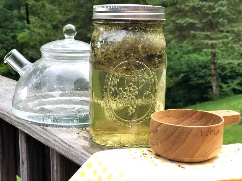 A glass tea kettle, mason jar filled with herbs and water, and wooden measuring cup are in the foreground. Trees are behind them.