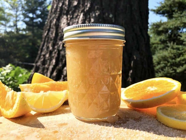 A jar of homemade coconut citrus sugar scrub is on a wooden cutting board with citrus fruits and spilled sugar. Trees are in the background.