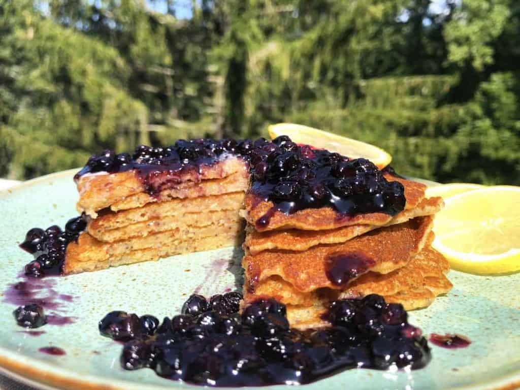 A stack of lemon poppy seed einkorn sourdough pancakes with blueberry syrup and lemons are in front of a pine tree in sunlight.