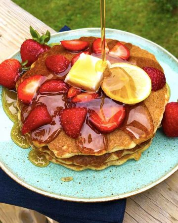 Maple syrup is being poured on a platter of lemon poppy seed pancakes with strawberries and vegan butter. The turquoise blue plate is on a navy blue linen napkin on a wood surface outdoors.