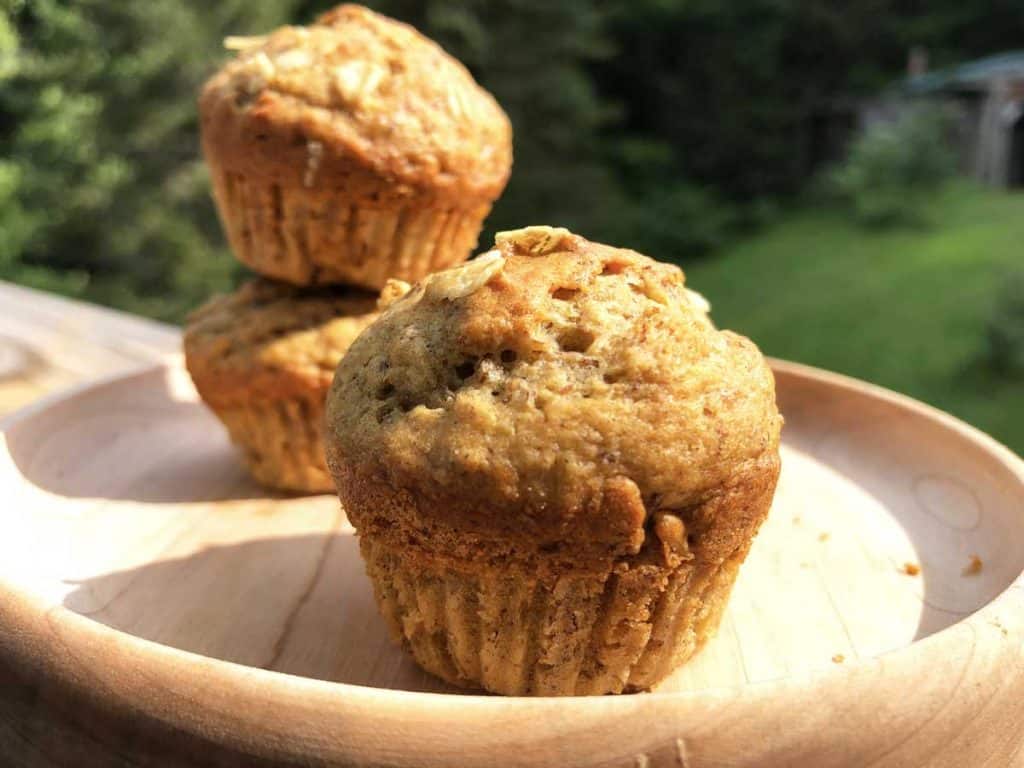 Three mini einkorn sourdough banana oat muffins are on a small maple plate. Trees and grass are in the background.