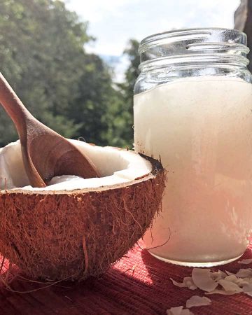 Half of a cracked coconut holds a wooden tablespoon. Beside it is a glass jar of coconut oil and coconut flakes. Sunlight is streaming in the photo.