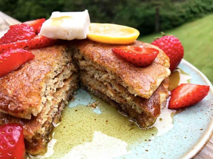 A closeup of lemon poppy seed einkorn sourdough pancakes with vegan butter, maple syrup, and sliced strawberries are on a blue plate outdoors. A large slice of the pancake stack has been eaten.