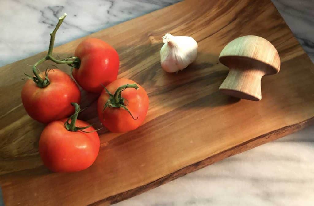 Four garden tomatoes on the vine, a head of garlic, and a wooden garlic masher are on a wooden cutting board on top of a marble surface.