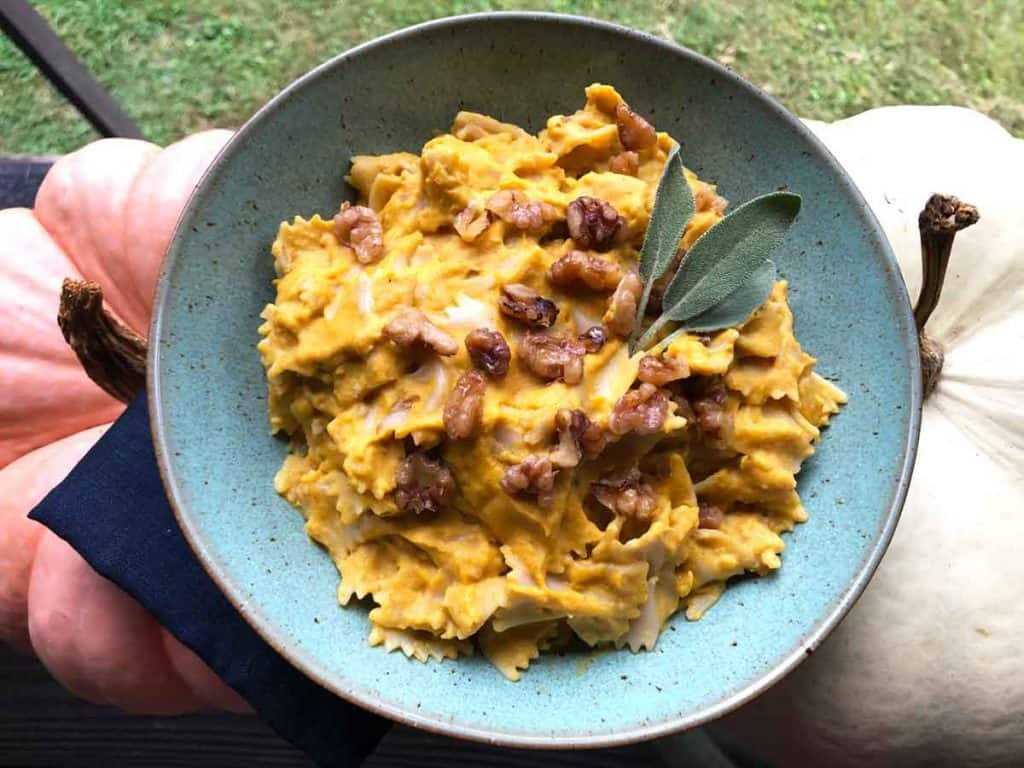 A bowl of pumpkin and sage pasta sauce on bow-tie pasta topped with toasted walnuts and fresh sage is atop a navy blue linen napkin and two pumpkins.