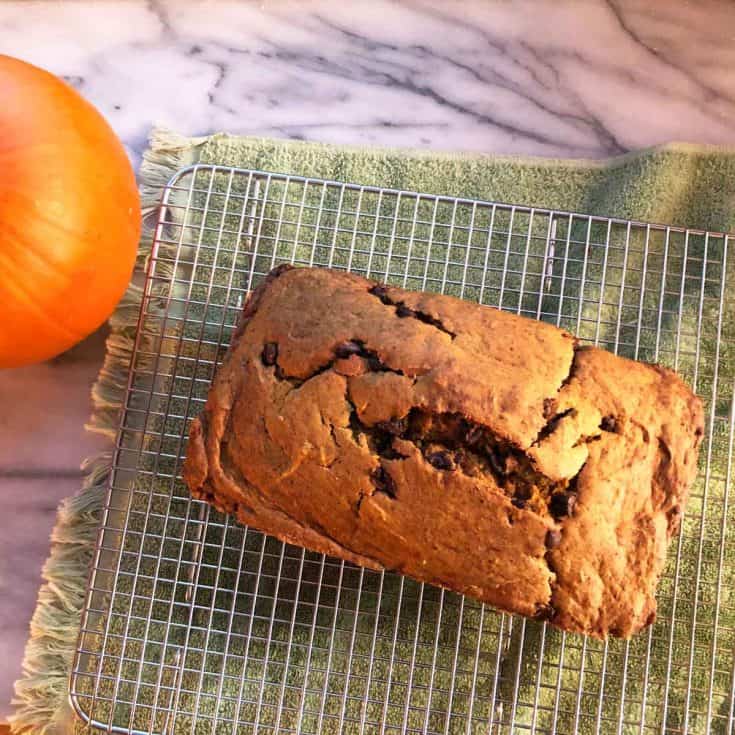 A loaf of einkorn pumpkin bread is on a cooling rack above a green towel on a marble surface. A pumpkin is nearby.