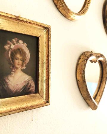 Several gold frames, mirrors, and a picture of a lady in a pink bonnet adorn a wall.