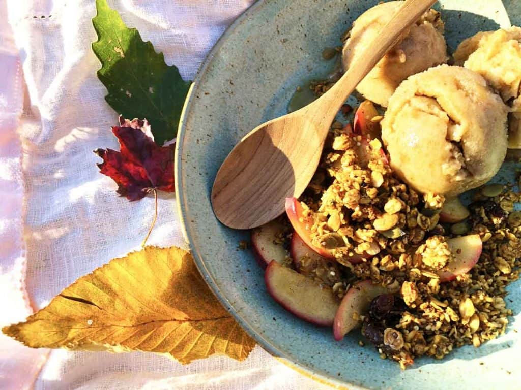 Three autumn leaves are beside a bowl with cinnamon apples, granola, and scoops of banana nice cream. They are on top of a pink linen place mat.