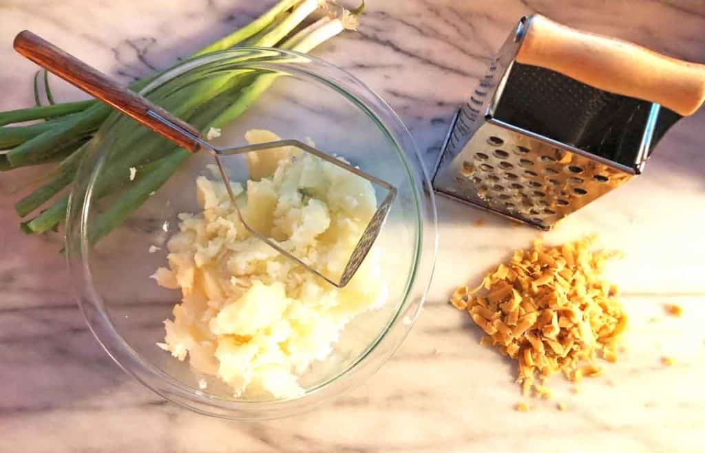 A glass bowl of mashed potatoes, a potato masher with a wooden handle, green onions, a cheese shredder, and shredded vegan cheese are on a marble countertop.