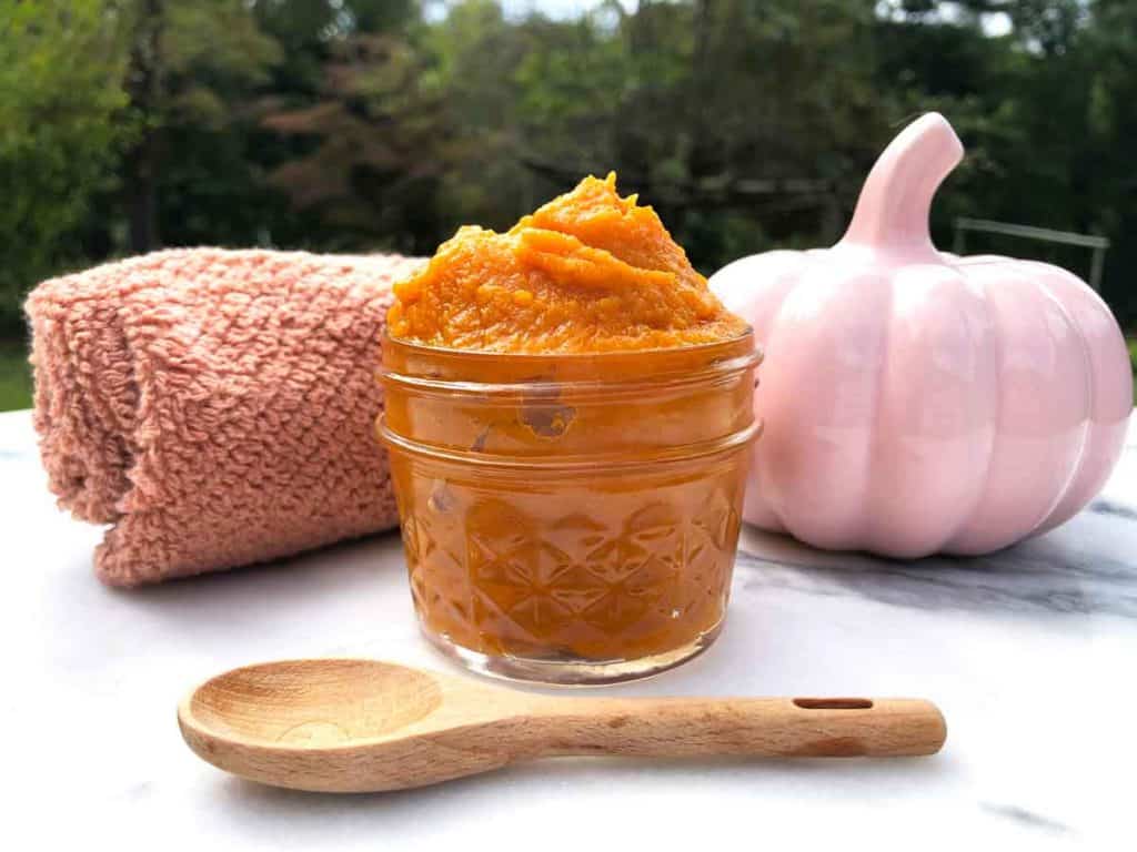 A wooden teaspoon is in front of a glass jar of pureed pumpkin, a decorative pink pumpkin, and a coral color washcloth outdoors on a marble table.
