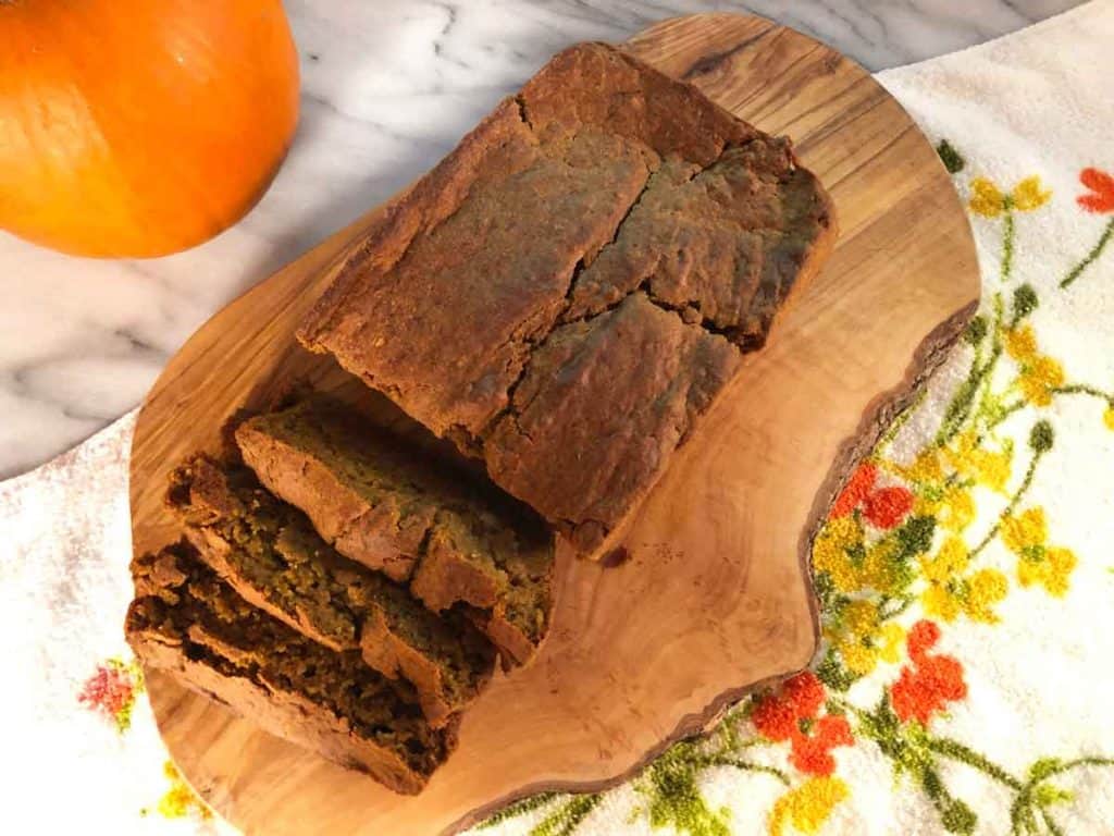 A loaf of einkorn pumpkin bread is sliced on a wooden cutting board atop a floral towel on a marble surface with a pumpkin.