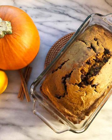 A loaf of einkorn pumpkin bread is cooling next to a pumpkin, an orange, and three sticks of cinnamon.