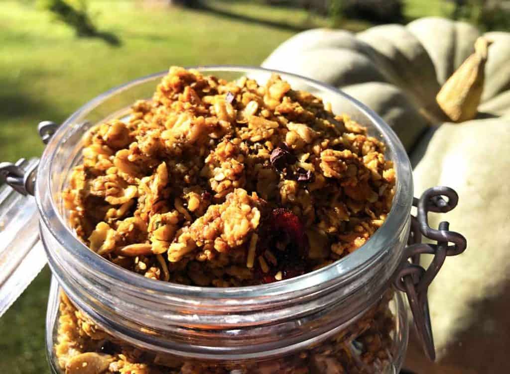 A large glass jar is full of pumpkin granola. It's in front of a green pumpkin outdoors.