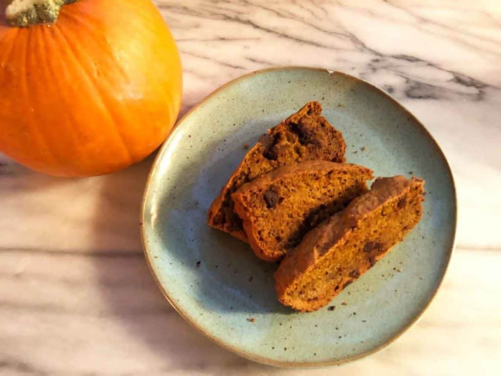 Three slices of einkorn pumpkin bread are on a blue plate next to a pumpkin on a marble surface.