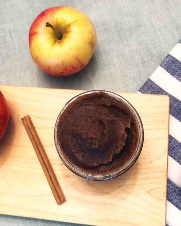 Apple butter in a small glass jar, a stick of cinnamon, and apples are on a wooden cutting board and a blue linen napkin and tablecloth.