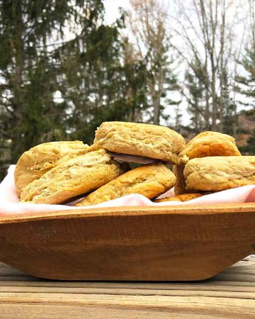 A linen lined dough bowl is filled with einkorn sourdough English muffins. It is outdoors in front of evergreen trees.