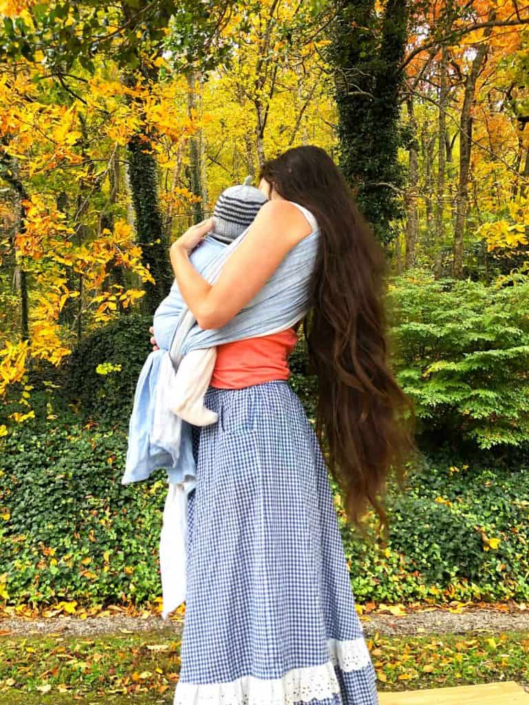 A mom in a long blue gingham skirt wears her baby in a woven wrap outdoors.