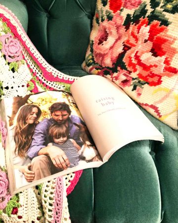 The Kind Mama book is opened and sitting on a green velvet armchair with a crocheted blanket and a needlepoint floral pillow.