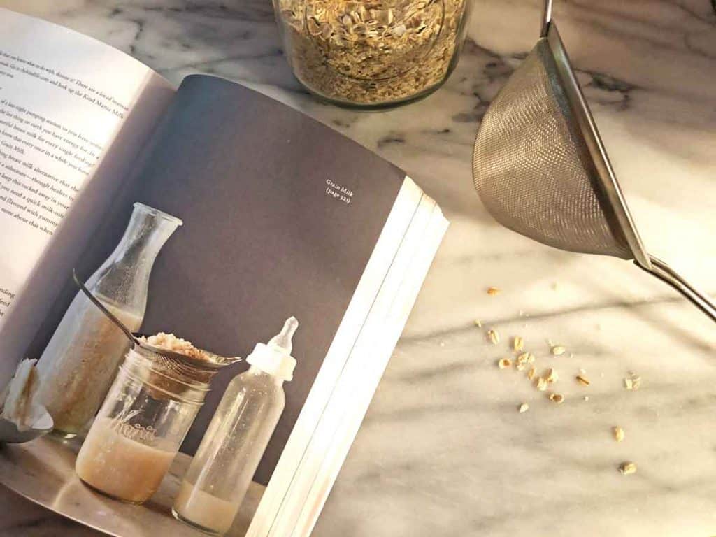 The Kind Mama book is opened to a page on baby's first food. It is on a marble surface with scattered oats and a stainless steel strainer.