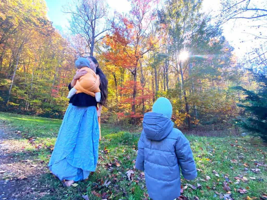 A woman has her infant in an orange woven wrap while her toddler is by her side in a coat. They are outdoors in autumn.