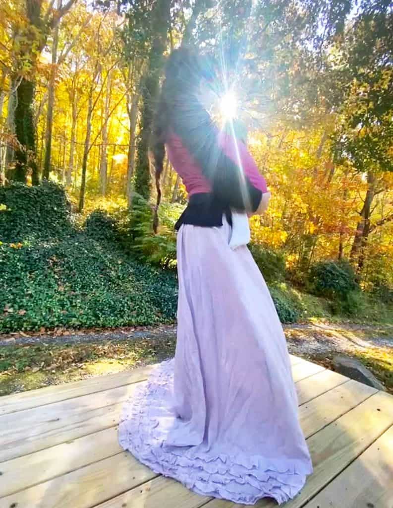 A woman with a long braided ponytail in a long purple skirt carries her baby in a pink woven wrap by a forest in autumn. Sunlight is shining through the leaves.