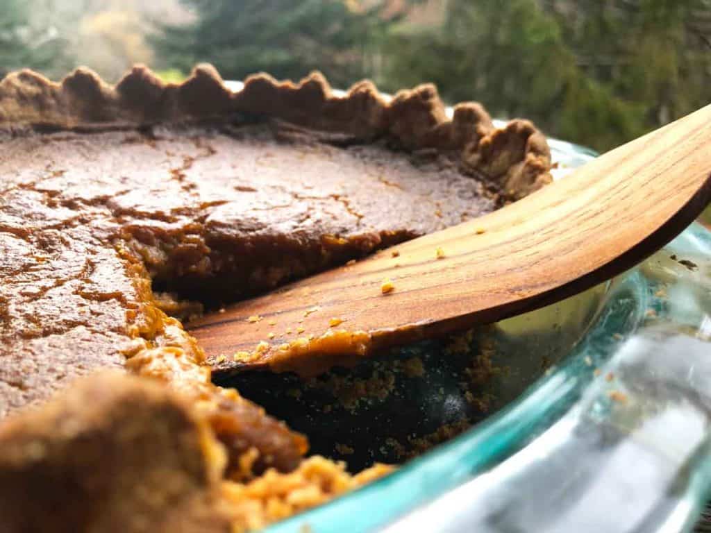 A plant-based pumpkin pie has been sliced. A wooden pie server is in the spot where a slice was. Trees are in the background.