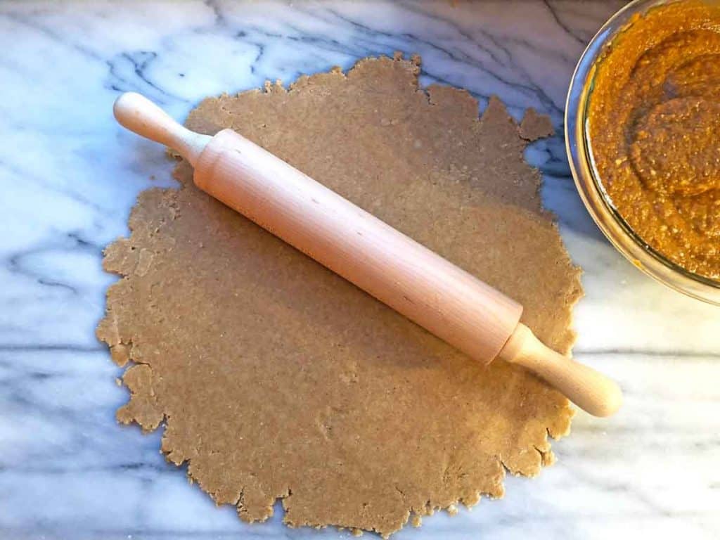 A wooden rolling pin rests on rolled out pie crust. They are on a marble surface beside a glass bowl of pumpkin pie filling.