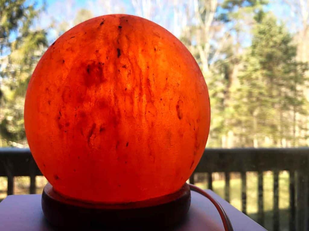 A salt lamp globe is by a window. A wooden railing, blue sky, and green trees are seen outdoors.