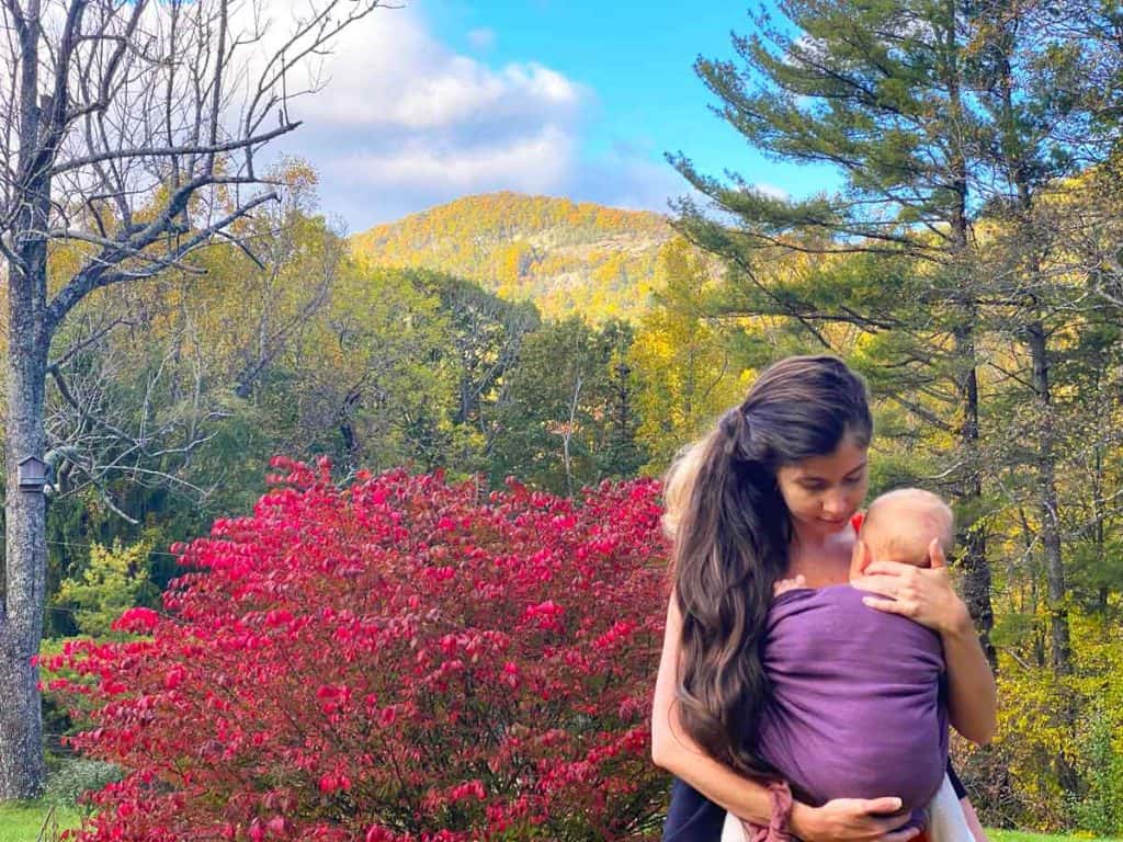 A mom wears two woven wraps: one for her toddler on her back and the other for her infant in front. Trees and mountains are in the background.