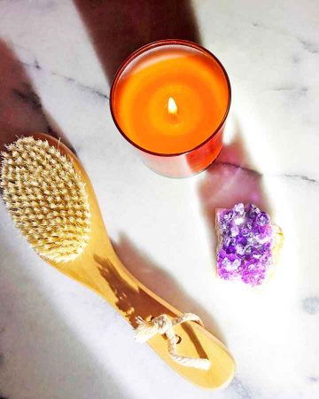 A dry body brush is beside a candle and an amethyst crystal on a marble table.