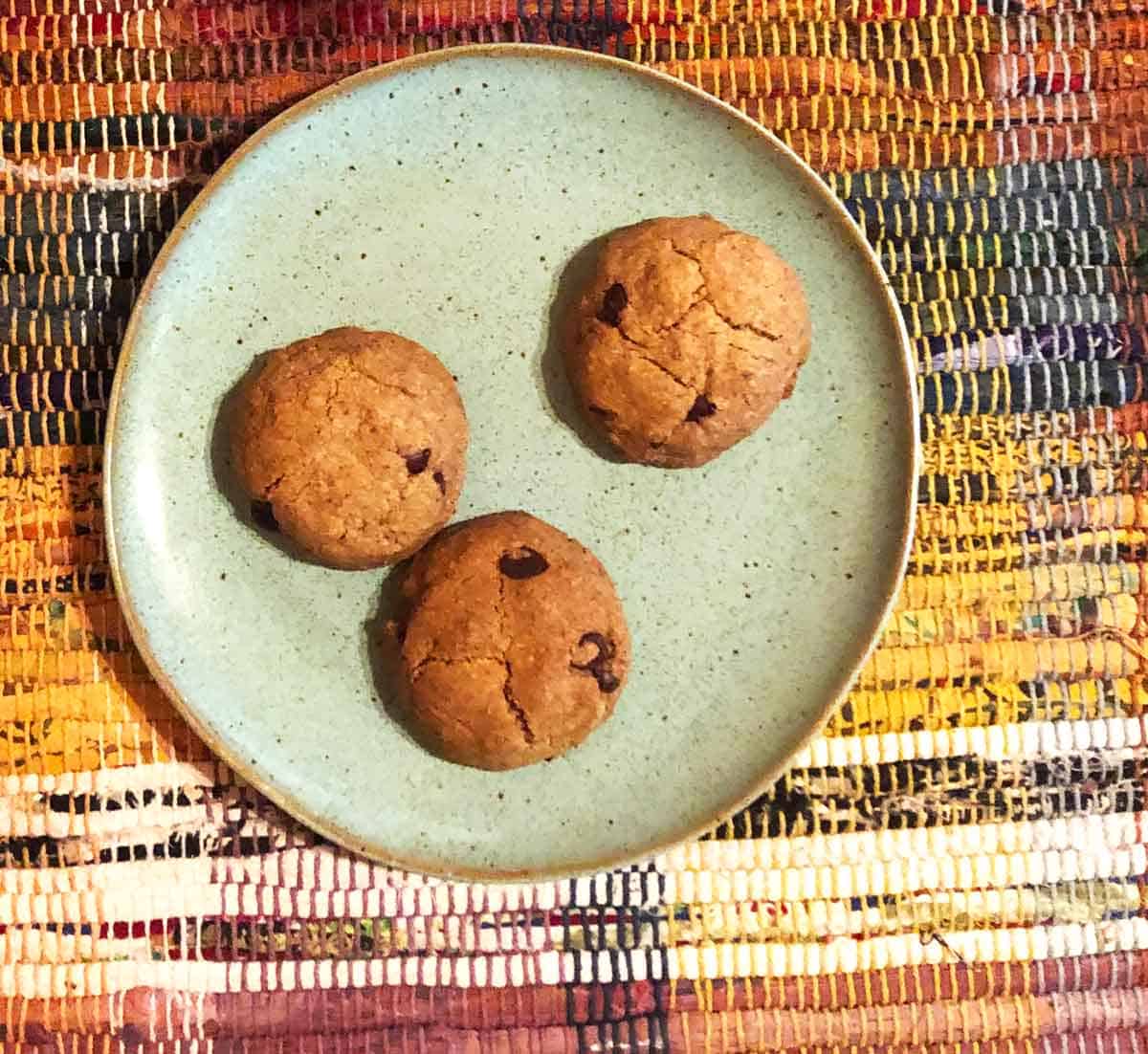 Three chocolate chip cookies are on a blue plate on top of a colorful plaid placemat.