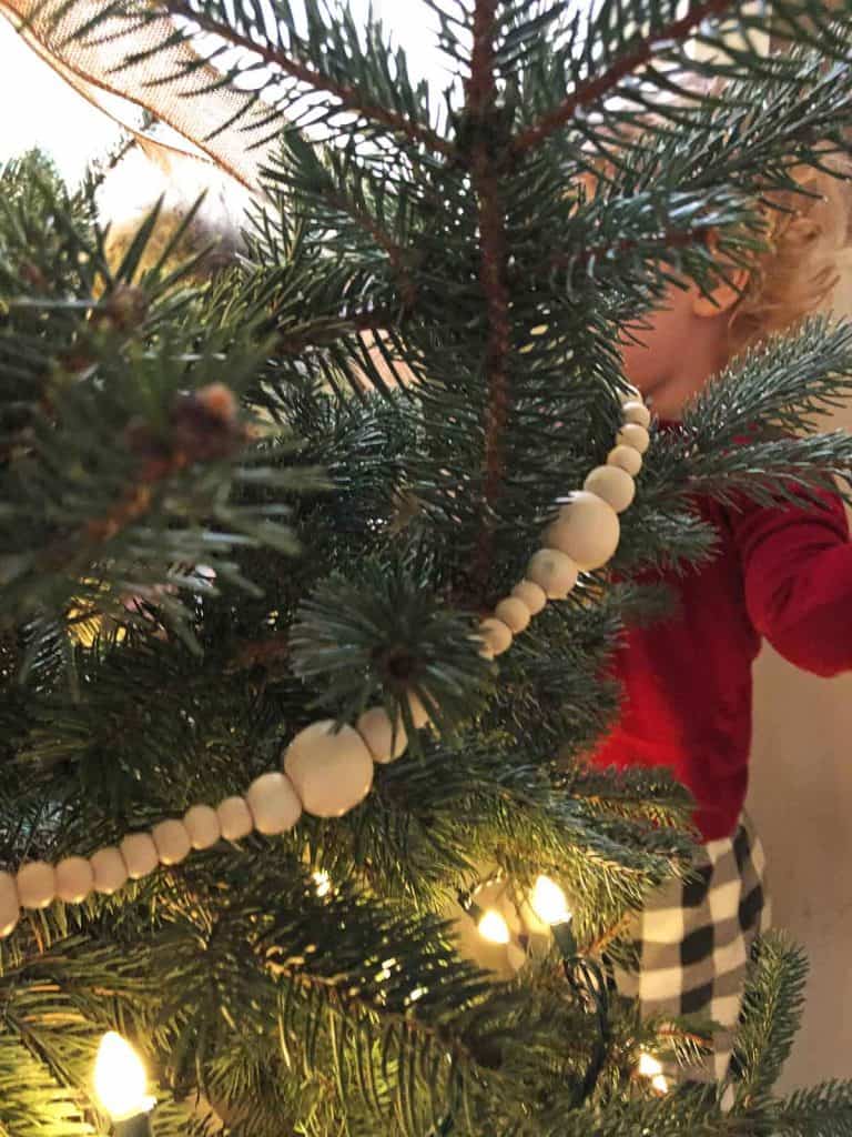 Wooden garland is seen on a live Christmas tree. A toddler decorates it in the background. 