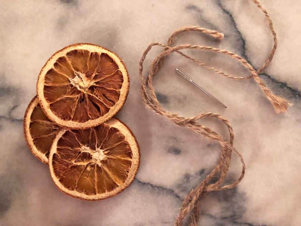 Three dried orange slices are on a marble surface beside twine and a sewing needle.