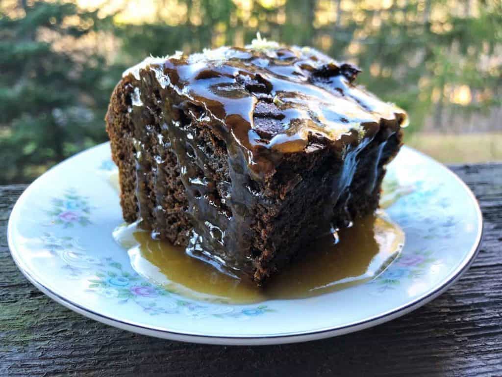 A slice of einkorn gingerbread is served with a lemon glaze. It is outdoors in front of evergreen trees. 
