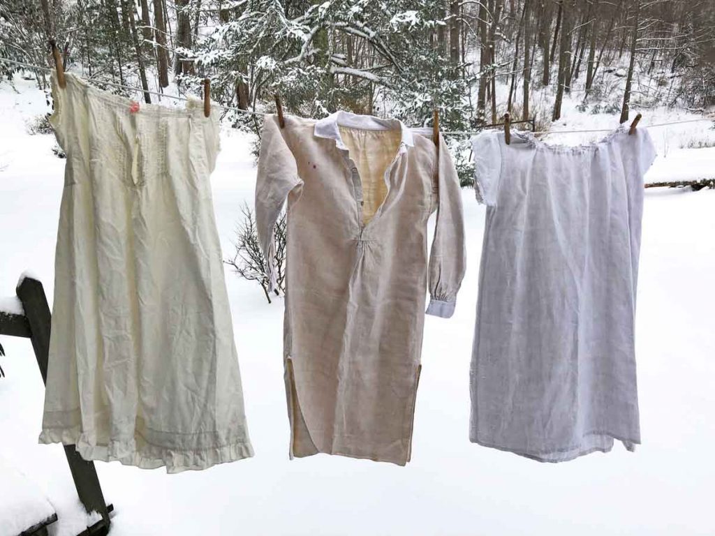 Three antique nightgowns are hanging on a clothesline in winter. Snow is on the ground and a forest is in the background.