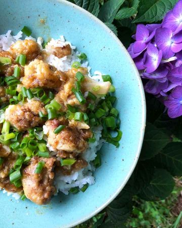 A bowl of vegan orange cauliflower is served with rice and green onions is beside a purple hydrangea.
