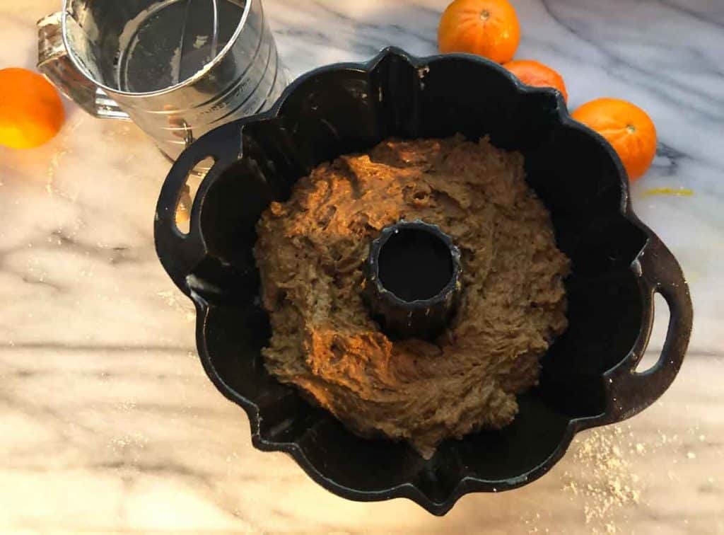Einkorn cake is in a cast iron bundt pan. Oranges and an old-fashioned sifter are beside it on a marble surface.
