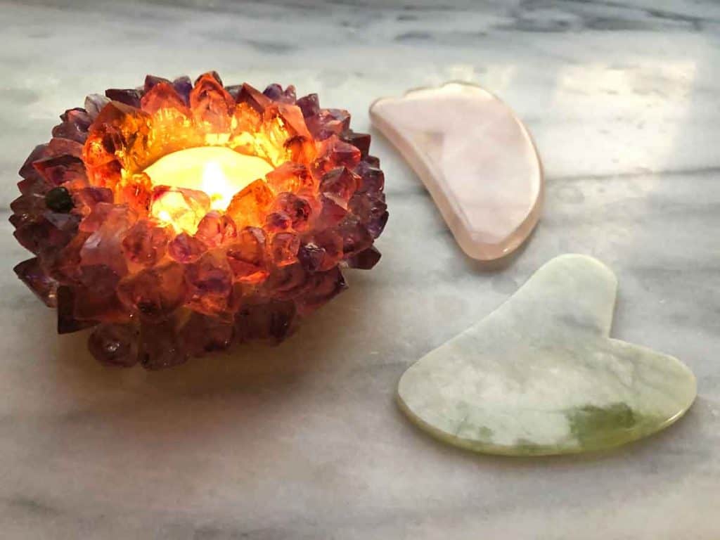 An amethyst candle holder with a lit tea light is beside two gua sha tools. One is rose quartz, the other is made from jade. 