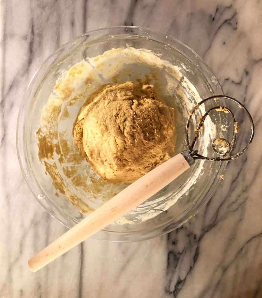 Einkorn sourdough is in a glass bowl on a marble surface. A wooden handled dough whisk rests on top of it. 