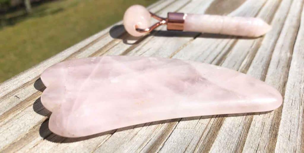 A rose quartz gua sha facial massage tool is in the foreground. In the background is a rose quartz face roller. Both are on a wooden surface outside.