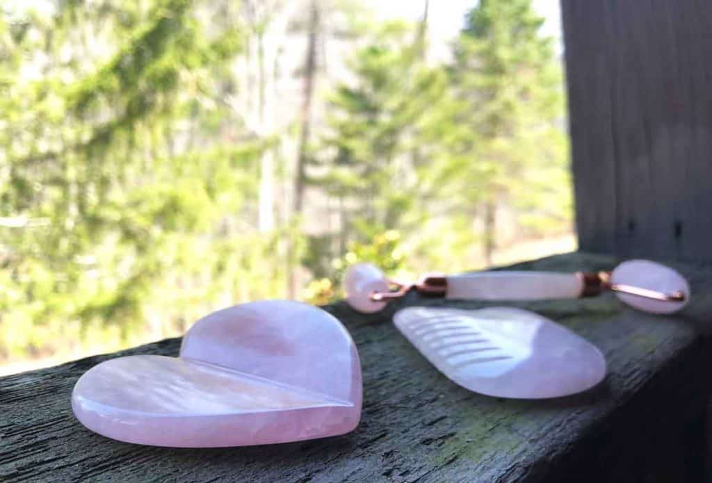 Three rose quartz beauty tools are outdoors on a wooden ledge in front of evergreen trees.