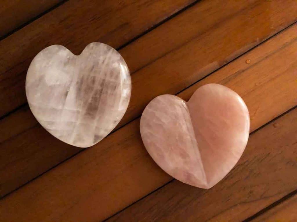 Two heart-shaped rose quartz gua sha tools are on a teak shower bench beside beads of water.