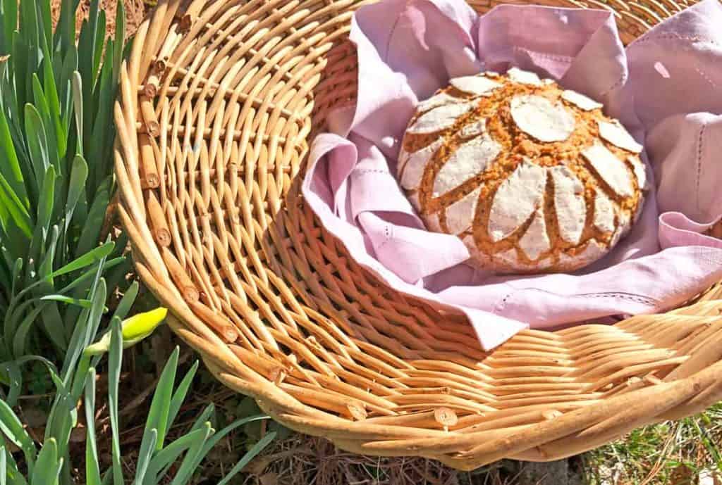 Einkorn sourdough bread scored with a sunflower design is in a lavender linen-lined basket beside of budding iris flowers. 