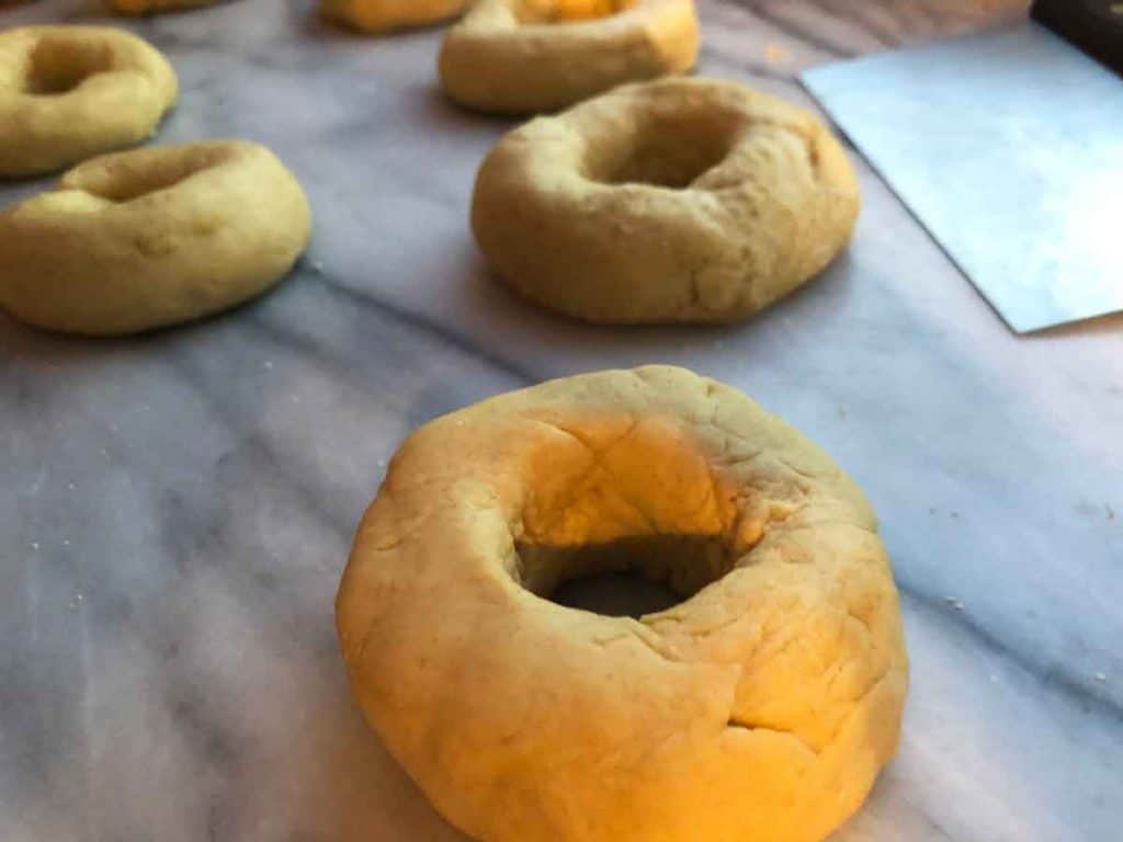 Einkorn sourdough bagels have been shaped and are resting on a marble surface beside a bench scraper.