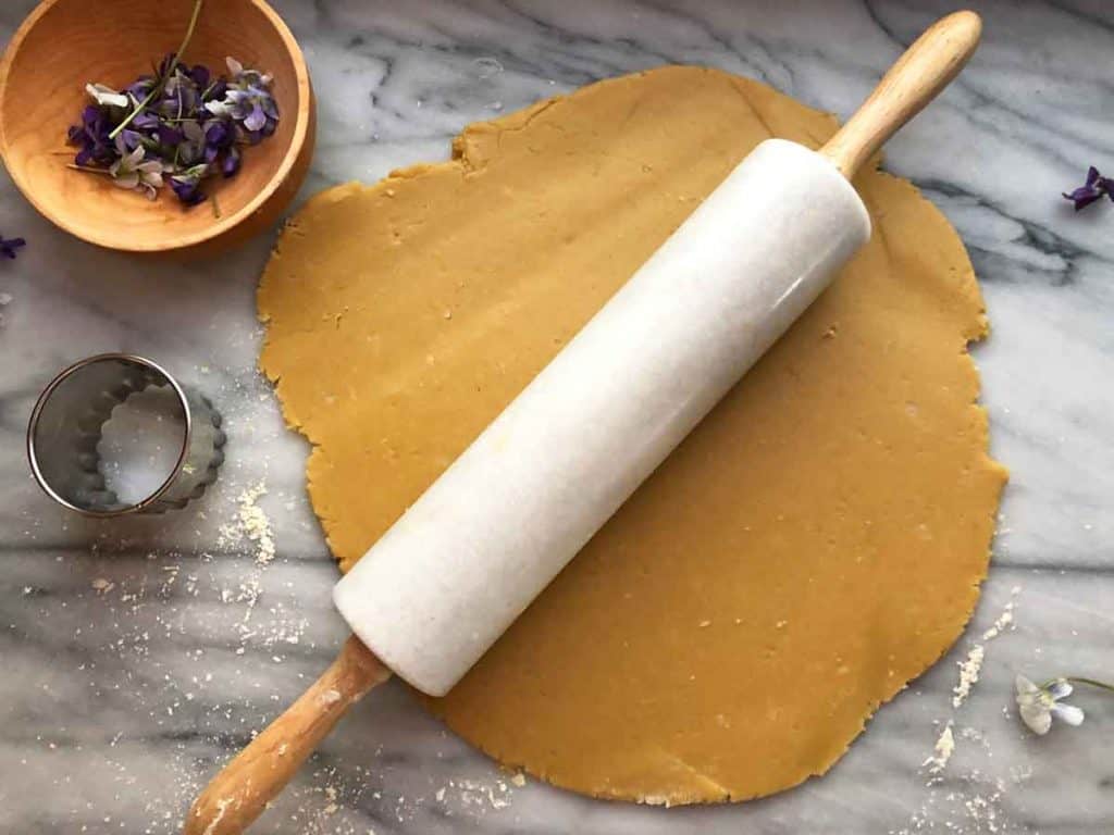 A marble rolling pin rolls out shortbread. A wooden bowl full of edible flowers and a fluted cookie cutter is beside the dough.