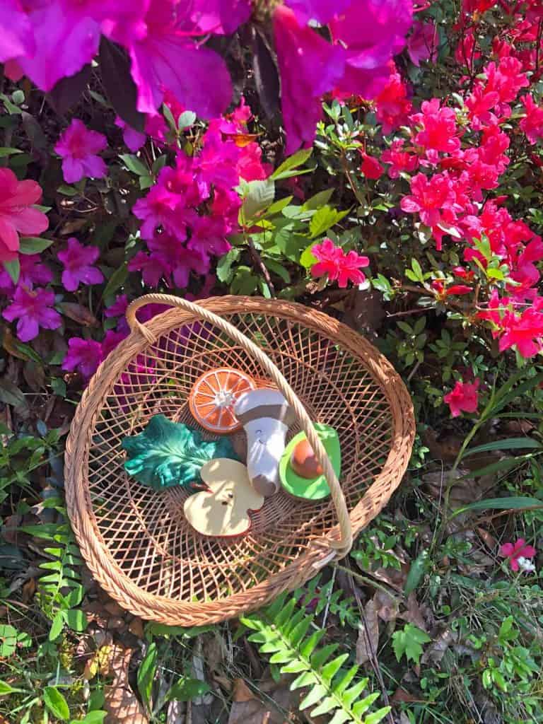 A woven basket is filled with natural teething toys and placed near blooming pink flowers.