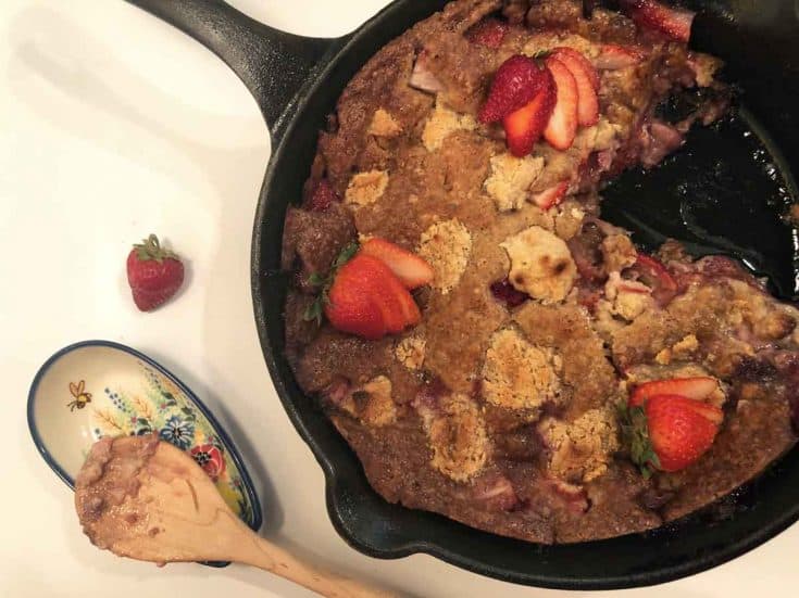 Einkorn strawberry cream cheese cobbler in a cast iron skillet beside a wooden spoon on a floral spoon rest.