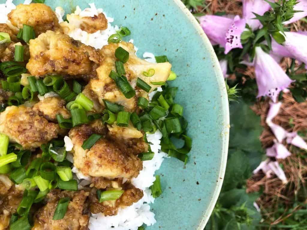 A bowl of orange cauliflower garnished with green onions is by foxglove flowers.