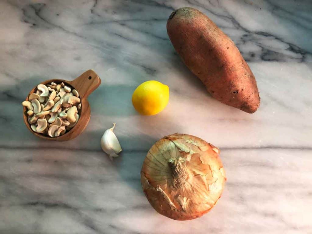 A wooden measuring cup with cashews is on a marble surface beside an unpeeled clove of garlic, a lemon, an onion, and a sweet potato.